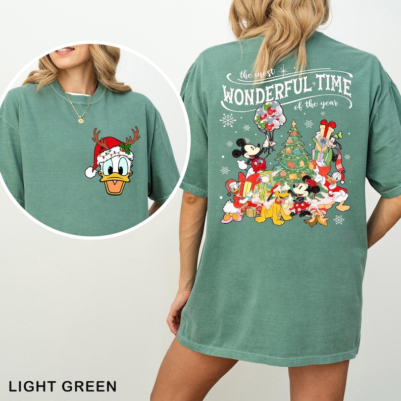 Puede incluir: Camiseta verde claro con un &aacute;rbol de Navidad y personajes de Mickey Mouse en la espalda. La parte delantera de la camiseta presenta una imagen de dibujos animados de Donald Duck con un gorro de Pap&aacute; Noel y cuernos de reno. El texto en la parte posterior dice "the most WONDERFUL TIME of the year".