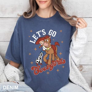 May include: A blue t-shirt featuring a cartoon cowgirl and horse graphic. The text "Let's Go Cowgirls" is printed on the shirt.