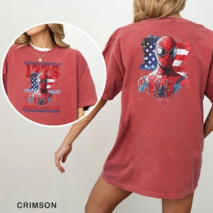 May include: A crimson short-sleeve t-shirt featuring a graphic of Spider-Man against a distressed American flag. The back displays a large print of the character, while the front shows a smaller version with the text 1776 UNITED STATES INDEPENDENCE DAY.