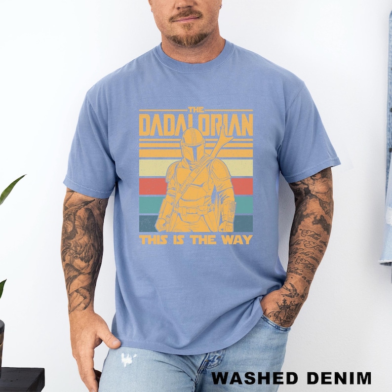 May include: A light blue t-shirt featuring a graphic of a Mandalorian-style character with the text "THE DADALORIAN" and "THIS IS THE WAY" in a retro design. The shirt is made of a soft material and has short sleeves.