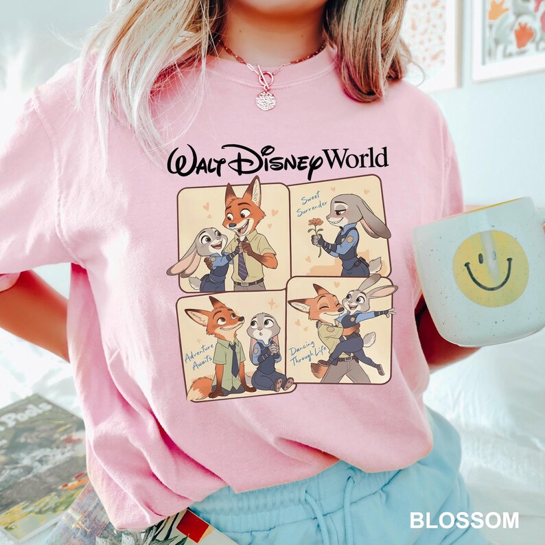 以下が含まれることがあります： ウォルト・ディズニー・ワールドのピンク色のTシャツ。映画「ズートピア」のニック・ワイルドとジュディ・ホップスの漫画のグラフィックが描かれています。グラフィックには、さまざまなポーズのキャラクターが描かれた4つのパネルが含まれています。黄色のスマイリーフェイスの白いマグカップが画像に写っています。