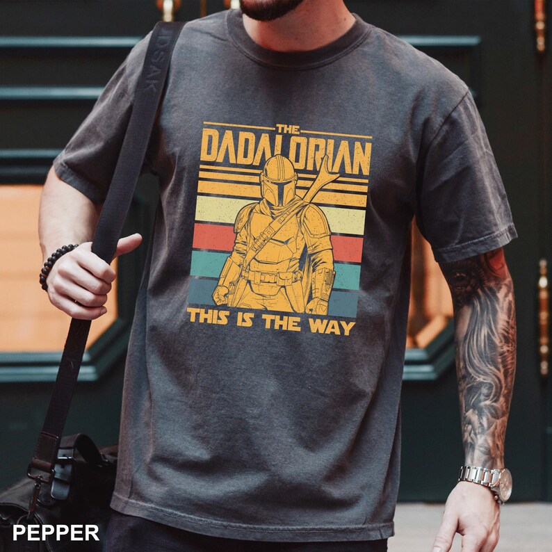 May include: Dark gray t-shirt featuring a retro design with the text "The Dadalorian" and an illustration of a Mandalorian-style character. Below the image is the text "This is the Way". The shirt has a vintage, distressed look.