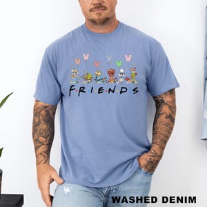 May include: A pale blue t-shirt featuring the word "FRIENDS" in black. Above the word are cartoon characters with balloons. The shirt is made of a soft material and has the words "WASHED DENIM" printed at the bottom.