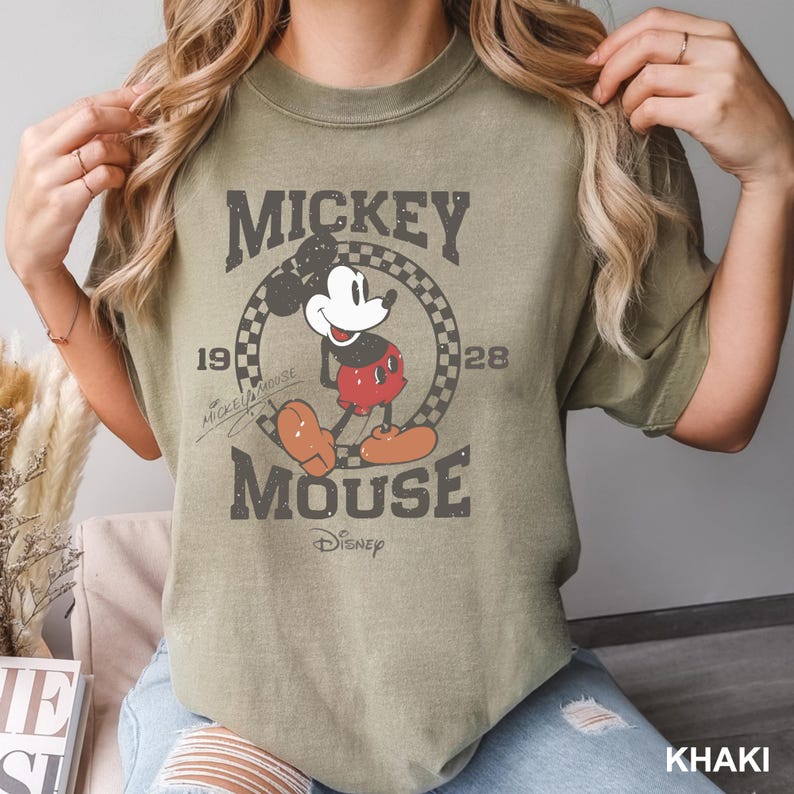 May include: A khaki green t-shirt with a vintage-style graphic of Mickey Mouse. The graphic features Mickey Mouse in a red and black outfit, with the text "Mickey Mouse" and "1928" around him. The text "Disney" is also included in the graphic.