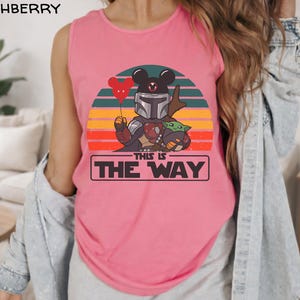 May include: Pink tank top featuring a graphic of a character in Mandalorian armor with a Mickey Mouse head, holding a red balloon. The design includes Baby Yoda and the text "THIS IS THE WAY" in a black rectangle, set against a sunset background.