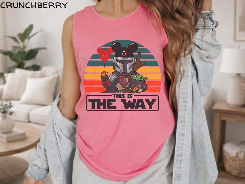 May include: Pink tank top featuring a graphic of a character in Mandalorian armor with a Mickey Mouse head, holding a red balloon. The design includes Baby Yoda and the text "THIS IS THE WAY" in a black rectangle, set against a sunset background.
