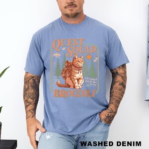 May include: Light blue t-shirt with a graphic of a cat wearing a backpack, the words "QUEST SQUAD" and "BROCCOLI" in orange, and the text "No One Wants It, But It's Good For You". The shirt is labeled "WASHED DENIM".