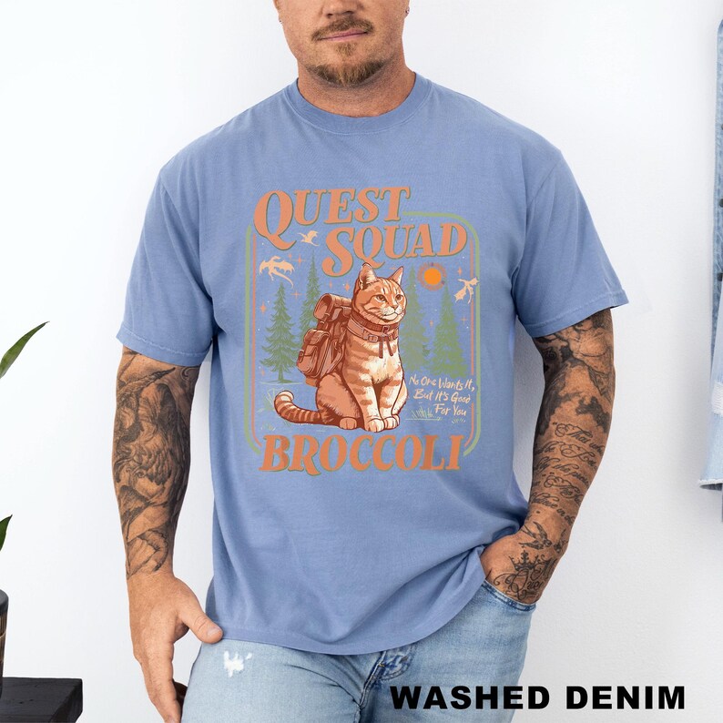 May include: Light blue t-shirt with a graphic of a cat wearing a backpack, the words "QUEST SQUAD" and "BROCCOLI" in orange, and the text "No One Wants It, But It's Good For You". The shirt is labeled "WASHED DENIM".