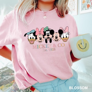 May include: Pink t-shirt with a graphic of Mickey Mouse and friends. The graphic includes Mickey Mouse, Minnie Mouse, Donald Duck, Goofy, and Pluto. The text below the graphic reads "Mickey & Co. Est. 1928".