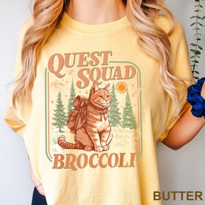 May include: Butter-colored t-shirt with a graphic featuring a cat wearing a backpack, the words "QUEST SQUAD" and "BROCCOLI". The design includes trees, a sun, and the text "No One Wants It But It's Good Stuff For You".