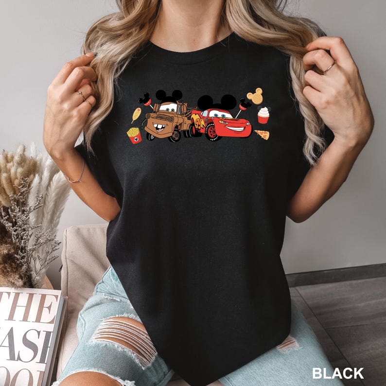 May include: Black t-shirt with a graphic of Lightning McQueen and Mater from Cars, wearing Mickey Mouse ears and surrounded by food items like fries, pizza, and a cupcake.