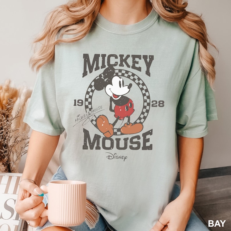 May include: A light blue t-shirt featuring a vintage-style graphic of Mickey Mouse. The graphic shows Mickey Mouse in a red and black outfit, standing in a checkered circle with the text "Mickey Mouse" and the years "1928" and "Mickey Mouse" written around the circle. The text "Disney" is written below the circle.