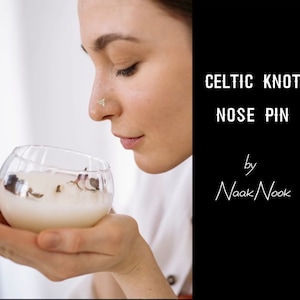 May include: A close-up of a person wearing a Celtic knot nose pin. The pin is made of gold and has a delicate design. The person is holding a glass bowl with a white substance inside.