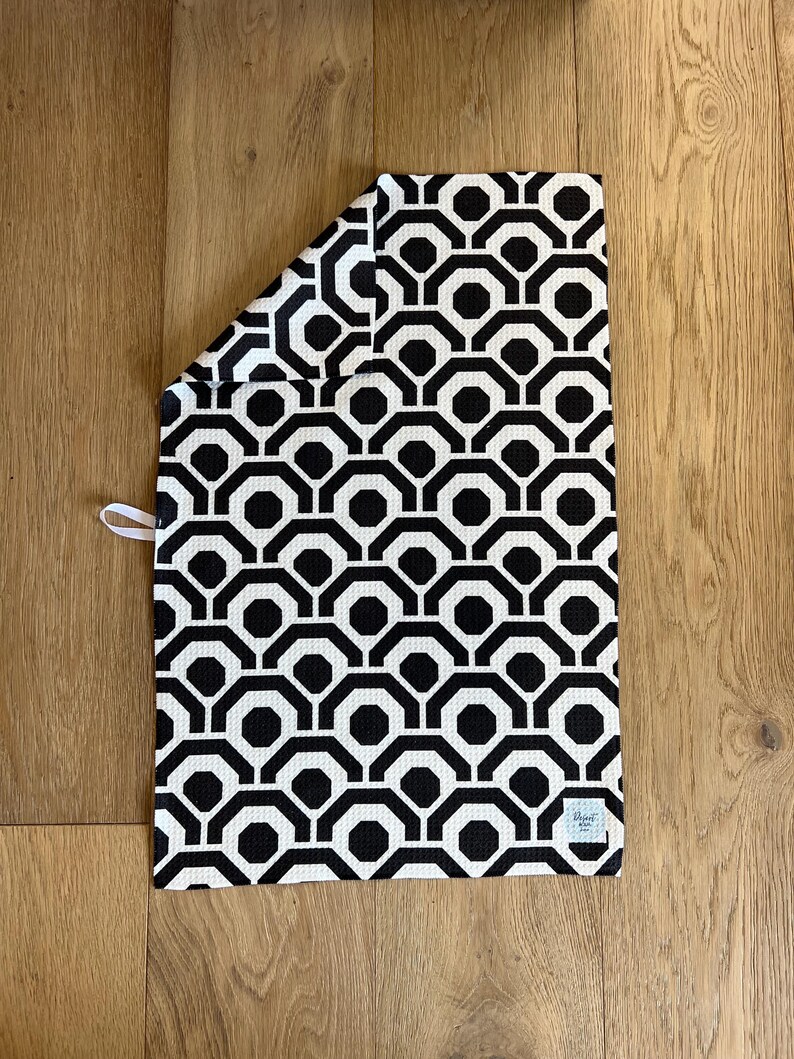 May include: Black and white kitchen towel with a geometric pattern. The towel has a black border and a white label with the text "Elegant Home" in black.
