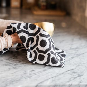 May include: A black and white geometric patterned kitchen towel is being used to dry a gold bowl on a white marble worktop.