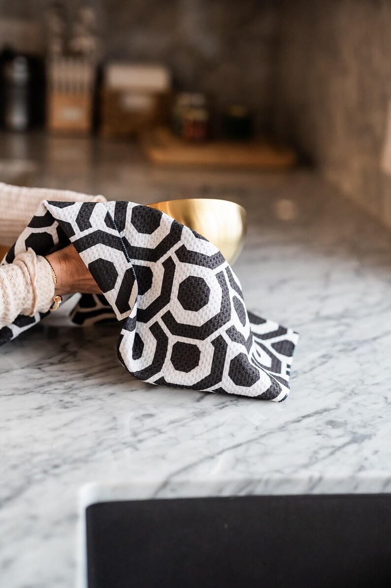 May include: A black and white geometric patterned kitchen towel is being used to dry a gold bowl on a white marble worktop.