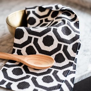 May include: A black and white geometric patterned kitchen towel with a wooden spoon on top. The towel is folded and sitting on a marble work surface.