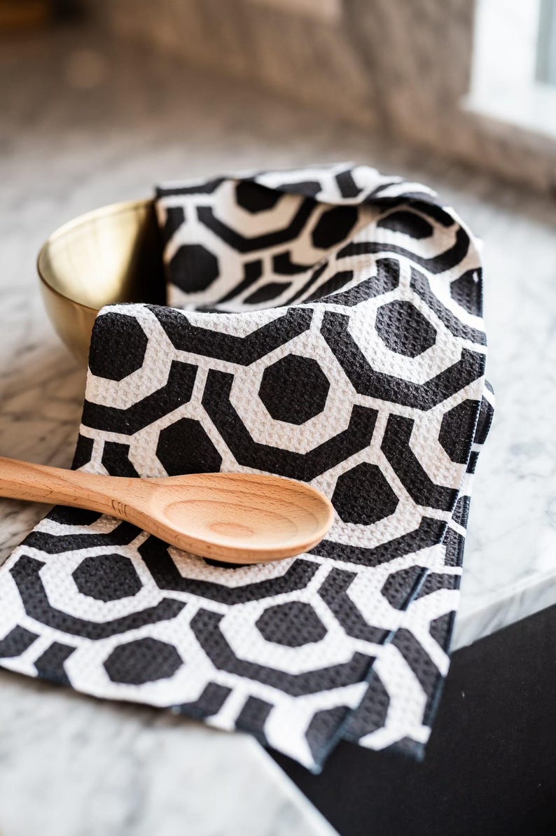 May include: A black and white geometric patterned kitchen towel with a wooden spoon on top. The towel is folded and sitting on a marble work surface.
