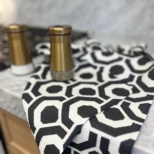 May include: A black and white geometric patterned kitchen towel with gold salt and pepper shakers on a marble worktop.