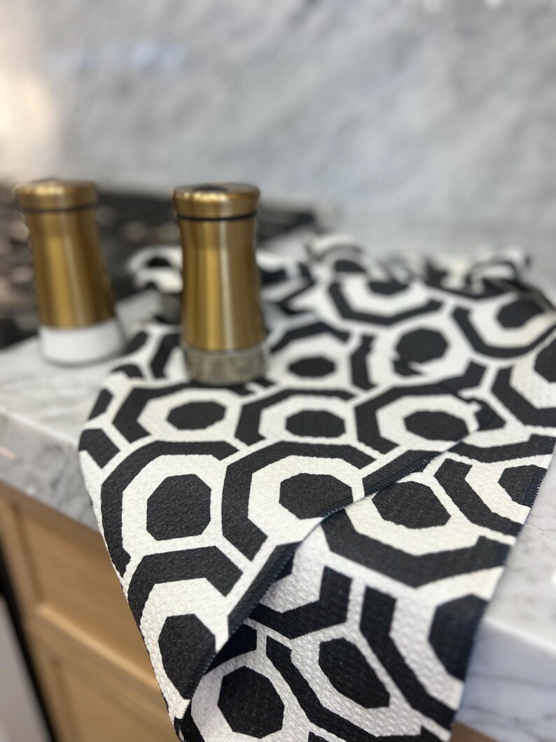 May include: A black and white geometric patterned kitchen towel with gold salt and pepper shakers on a marble worktop.