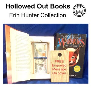 K&ouml;nnte beinhalten: Ein ausgeh&ouml;hltes Buch aus der Erin Hunter Collection, mit einem versteckten Fach, das Bargeld enth&auml;lt, das mit einem blauen Band gesichert ist. Ein Holzanh&auml;nger bietet eine kostenlose Gravur. Das Buch "Warriors: The Last Hope" ist ebenfalls sichtbar.