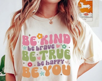 May include: A white t-shirt with a retro-style graphic that says "Be Kind, Be Brave, Be True, Be Happy, Be You" with colorful flowers and stars.