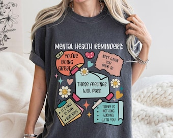 May include: A dark gray t-shirt with a graphic print of colorful sticky notes with positive affirmations about mental health. The text on the notes includes "You're doing great", "Rest when you need to", "These feelings will pass", and "It's okay to have bad days".