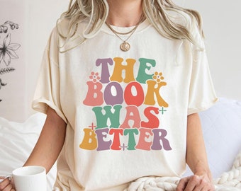 Może przedstawiać: Biały T-shirt z napisem "THE BOOK WAS BETTER" w kolorowych, retro literach. Litery są obwiedzione czarną ramką i mają lekko wypukły efekt.