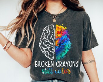 May include: A dark grey t-shirt with a graphic of a brain with one half in black and white and the other half in colorful watercolor. The text "BROKEN CRAYONS still color" is printed below the graphic.