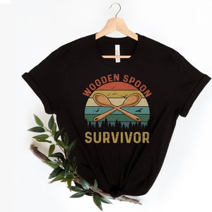 May include: Black t-shirt featuring a vintage-style graphic of two wooden spoons crossed over a sunset with the text "Wooden Spoon Survivor".