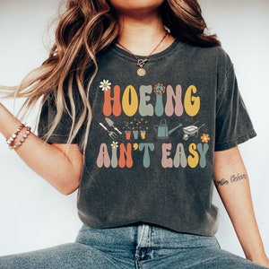 Op de afbeelding: Een donkergrijs T-shirt met een retro-grafische afbeelding met de tekst "HOEIN' AINT' EASY" in kleurrijke letters. De afbeelding bevat ook een vredesteken, een bloem en tuingereedschap.