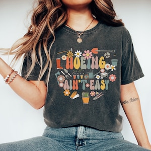 Op de afbeelding: Een donkergrijs T-shirt met een grafisch ontwerp van tuingereedschap en bloemen. De tekst op het shirt luidt "HOEING AIN'T EASY".