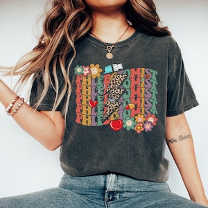 Puede incluir: Una camiseta gris oscuro con un diseño gráfico colorido que presenta la palabra "Phlebotomist" repetida varias veces, un rayo con estampado de leopardo, una manzana roja y flores.