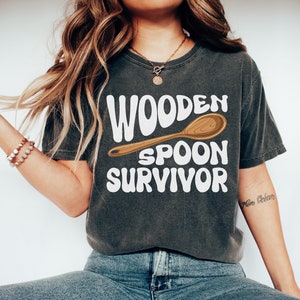 May include: A dark grey t-shirt with the text "Wooden Spoon Survivor" in white, featuring a cartoon wooden spoon.