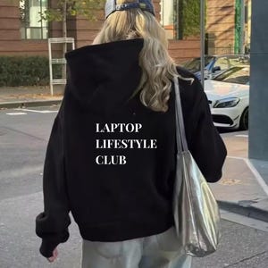 Könnte beinhalten: Ein schwarzer Kapuzenpullover mit dem weißen Text "LAPTOP LIFESTYLE CLUB". Die Person trägt eine blaue Baseballkappe und eine silberne Tragetasche. Die Person geht auf einer Stadtstraße.