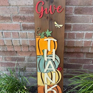 May include: Wooden Thanksgiving sign with the words "Give Thanks" and pumpkin designs. The sign is made of dark brown wood and features orange, teal, and yellow pumpkins. A red dragonfly and the word "Give" are also included.