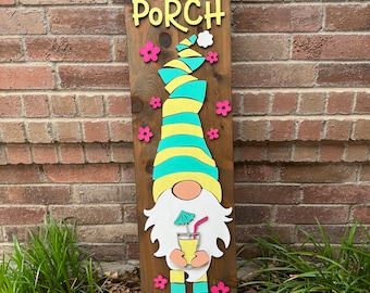 Puede incluir: Un letrero de bienvenida a nuestro porche con un gnomo con una bebida amarilla y un sombrero azul y amarillo. El letrero está hecho de madera y tiene flores rosas y una nube blanca.