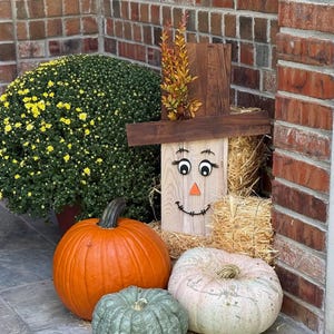 May include: A fall-themed outdoor display featuring a wooden scarecrow with a painted face, a straw hat, and a straw bale base. The display includes an orange pumpkin, a white pumpkin, a green pumpkin, and a large green bush with yellow flowers.