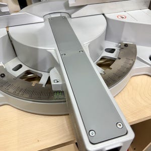 May include: A gray and silver miter saw with a long, rectangular gray guide. The saw has a circular blade guard and a measurement scale. The saw is on a light wood surface.