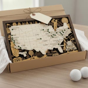 May include: A wooden wall art piece featuring a map of the United States with golf-themed cutouts including clubs, balls, and a cart, housed in a cardboard gift box with a blank tag. Two white golf balls and a wooden tee sit in the foreground.