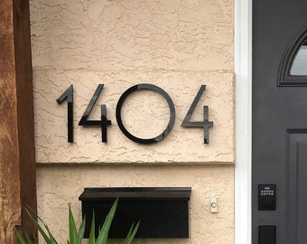 May include: A black house number plaque with the number 1404 on a light beige wall. A black mailbox is below the house number. A black door with a glass window is to the right of the house number. A potted plant is to the left of the house number.