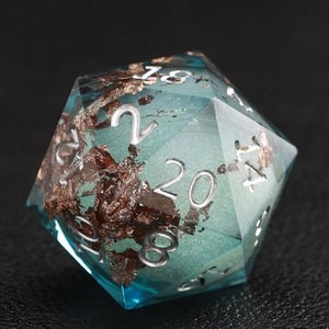 May include: A multi-sided, clear resin die with a teal interior and brown inclusions. The die has white numbering and is designed for tabletop gaming. The die is set against a dark background.