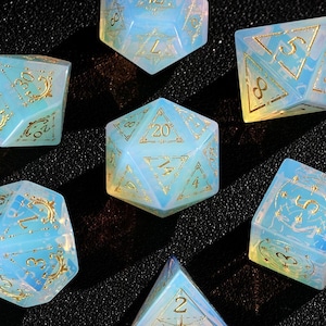 May include: Set of seven opalescent dice with gold numbering and accents. The dice are in various geometric shapes, including a d4, d6, d8, d10, d12, and d20. The dice have a light blue and white color scheme, with a shimmering effect.