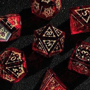 May include: Set of seven red and gold polyhedral dice for tabletop games. Each die features gold numbering and intricate designs. The dice are made of a translucent material, allowing light to pass through.