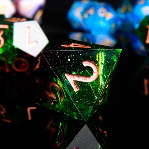 May include: A green and black iridescent dice with a copper coloured number two on it. The dice is a d6, or six-sided die.