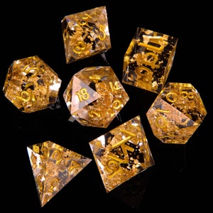 Puede incluir: Conjunto de siete dados transparentes con motas doradas y números dorados. Los dados son un d4, d6, d8, d10, d%, d12 y d20.