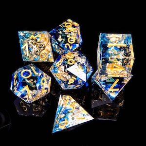 Könnte beinhalten: Ein Satz von sechs blauen und goldenen Würfeln mit goldenen Blatt-Akzenten. Die Würfel sind aus klarem Harz gefertigt und haben verschiedene Formen, darunter ein W4, W6, W8, W10, W12 und W20.