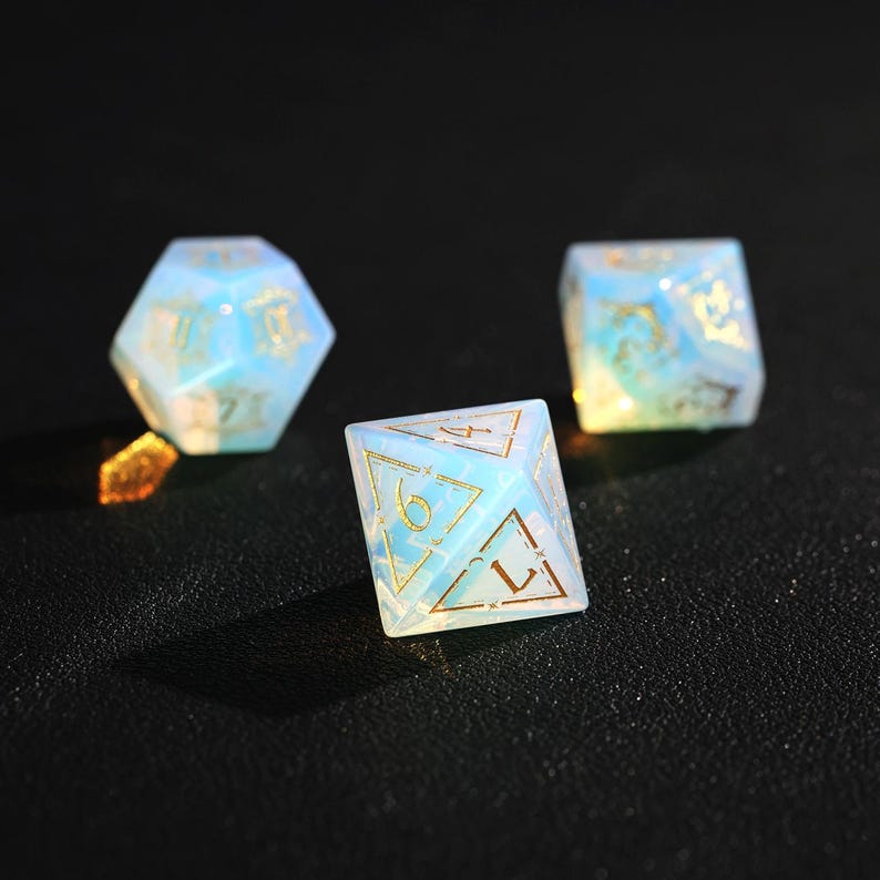 May include: Three opalescent dice with gold numbering and accents are displayed against a dark background. The dice are in various geometric shapes, including a 20-sided die, a cube, and a 8-sided die. The dice have a translucent, glowing appearance.