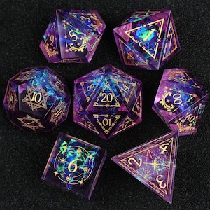 Puede incluir: Un juego de seis dados poliédricos hechos de resina púrpura translúcida con escamas de lámina iridiscente azul y verde incrustadas. Cada dado presenta números en tinta dorada y patrones de líneas geométricas decorativas en cada cara.