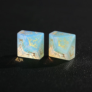 May include: Two opalescent dice with gold-colored filigree and numbers. The dice are a light blue color with a pearlescent sheen. The numbers are in a gold color. The dice are for a tabletop game.